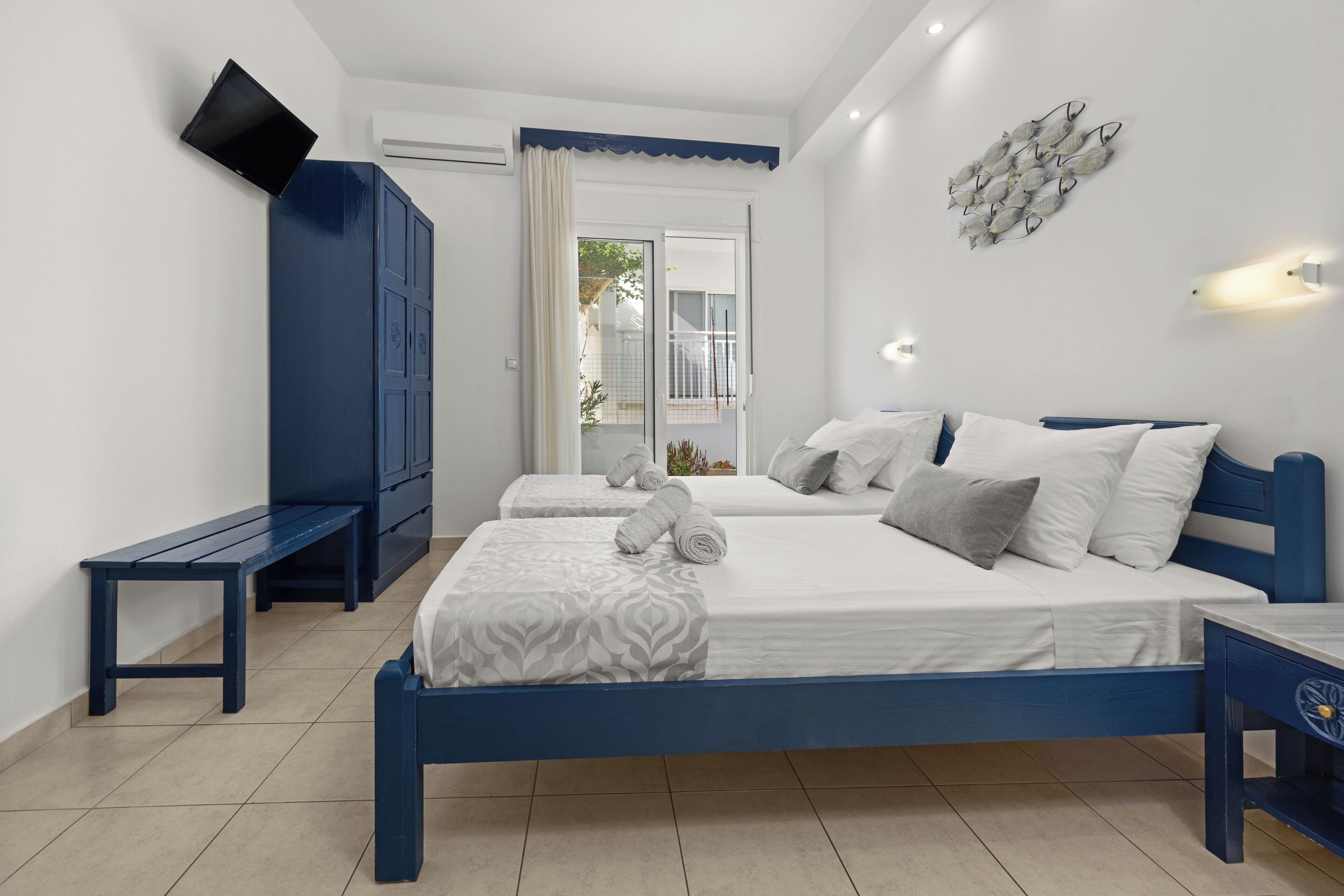 A bedroom with two beds, a bench, a TV, and a wall-mounted light fixture.
