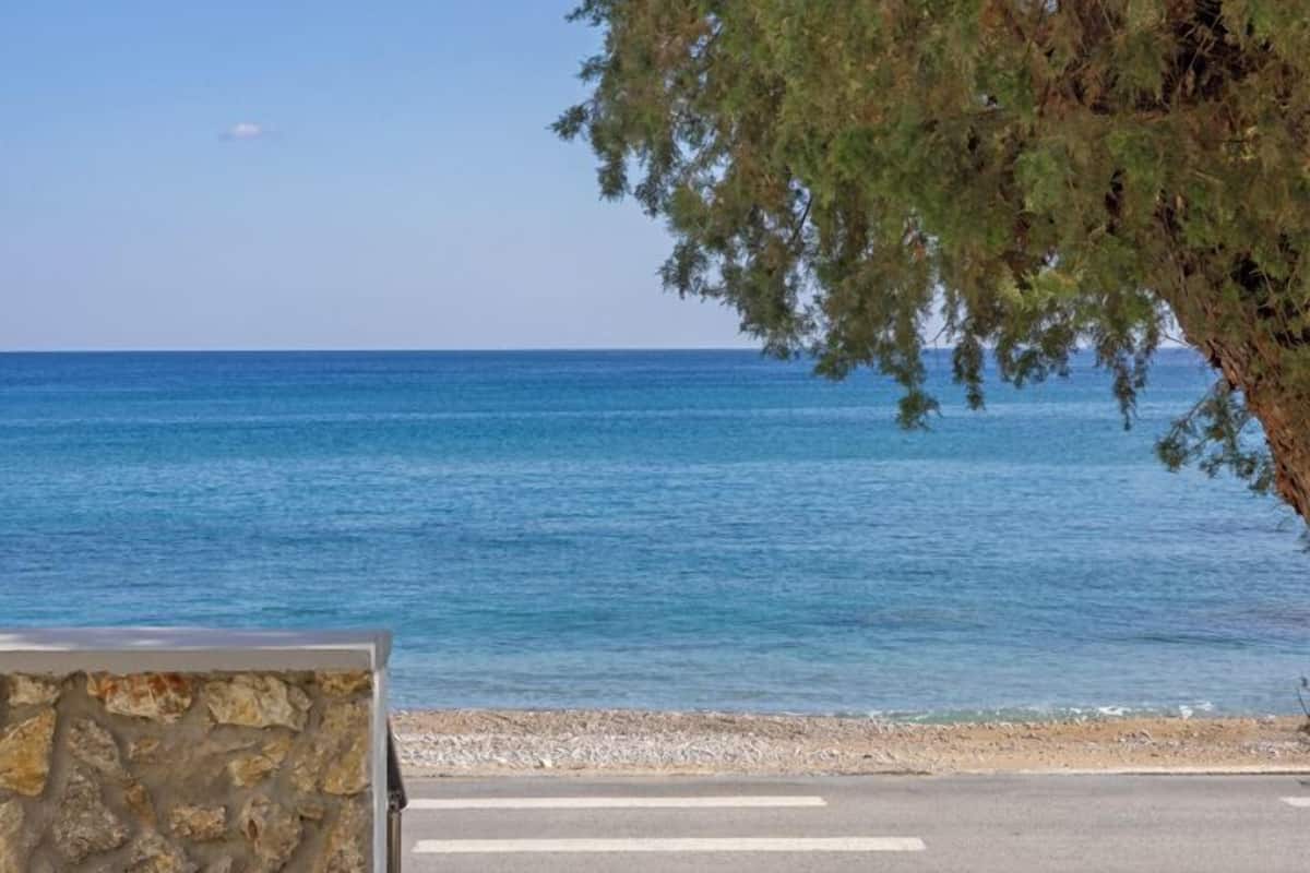 Vista costera con cielo despejado, playa de guijarros y un muro de piedra que conduce a una zona pavimentada.