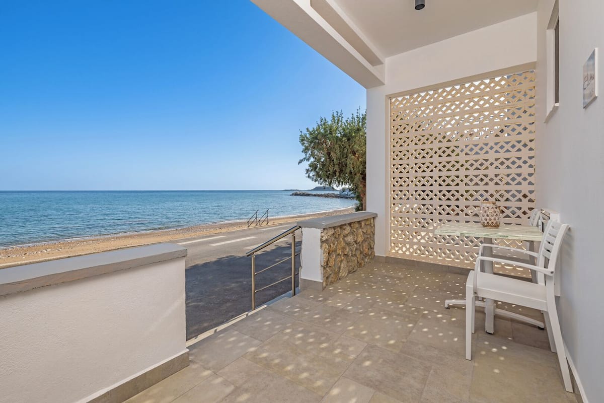 Un balcón con vista al mar, un muro de piedra, una mesa de vidrio con sillas y un enrejado decorativo.