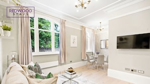 A modern living room with a sofa, a glass coffee table, and a dining area with a TV mounted on the wall. - THE FORBURY  (Reading)