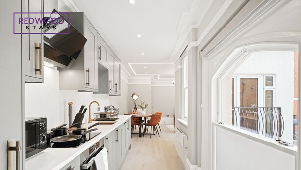 A modern kitchen with white cabinetry, a dining area with orange chairs, and a large window providing a view of the outside. - THE FORBURY  (Reading)