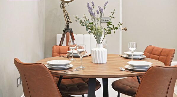 Een eettafel gedekt met borden, glazen en bestek, omringd door beklede stoelen in bruin. Op de tafel staat een witte vaas met lavendel en een ronde tafellamp.