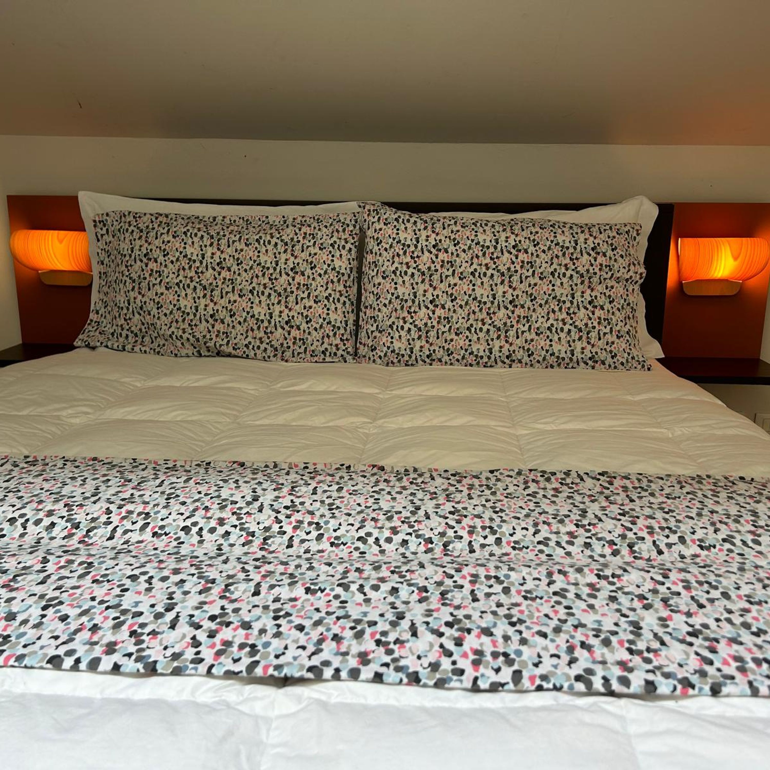 Habitación de hotel con una cama, dos almohadas y dos lámparas fijadas a la pared.