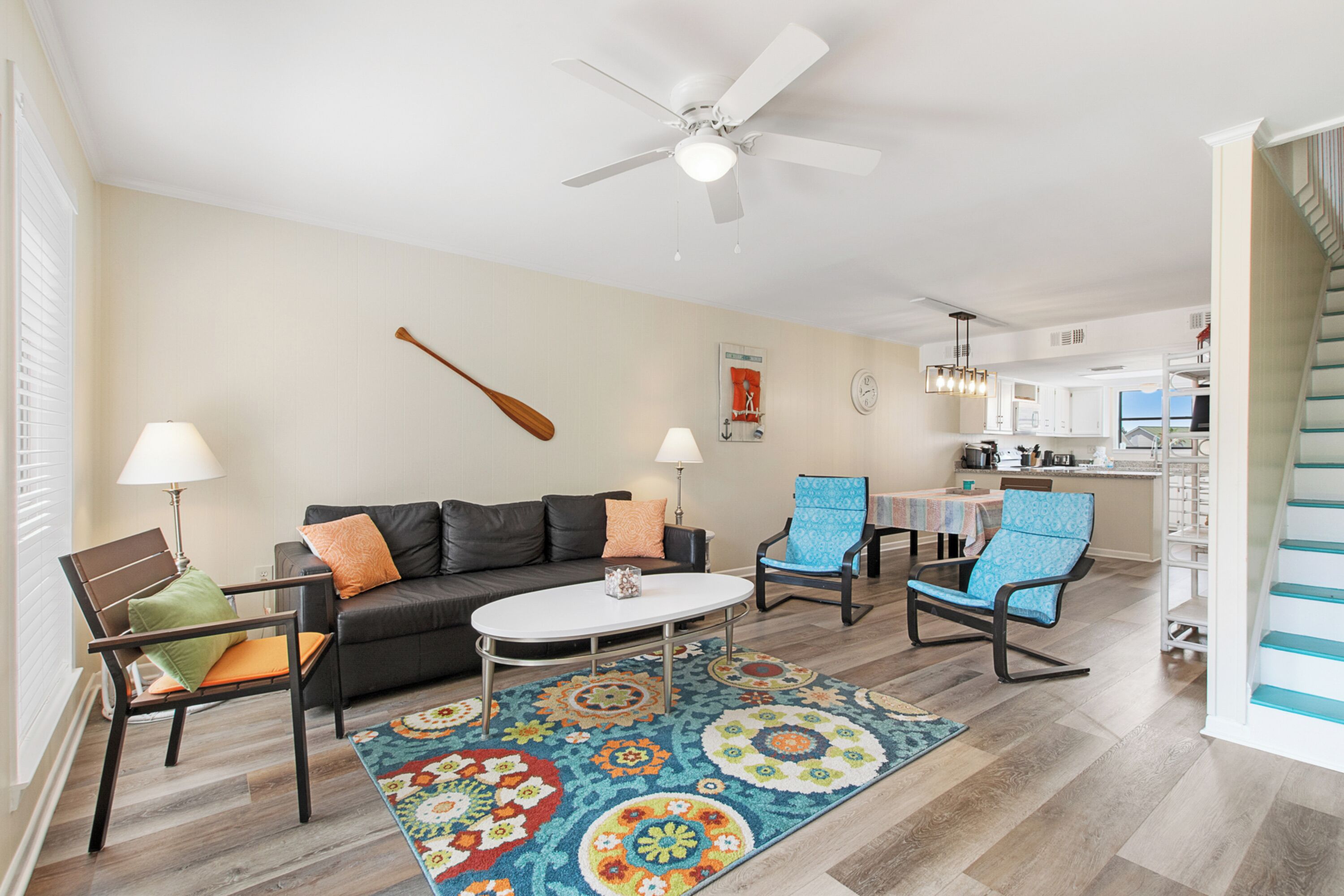 A living room with a sofa, two chairs, a coffee table, and a patterned rug. There is a ceiling fan, a wall-mounted guitar, and a kitchen area in the background.