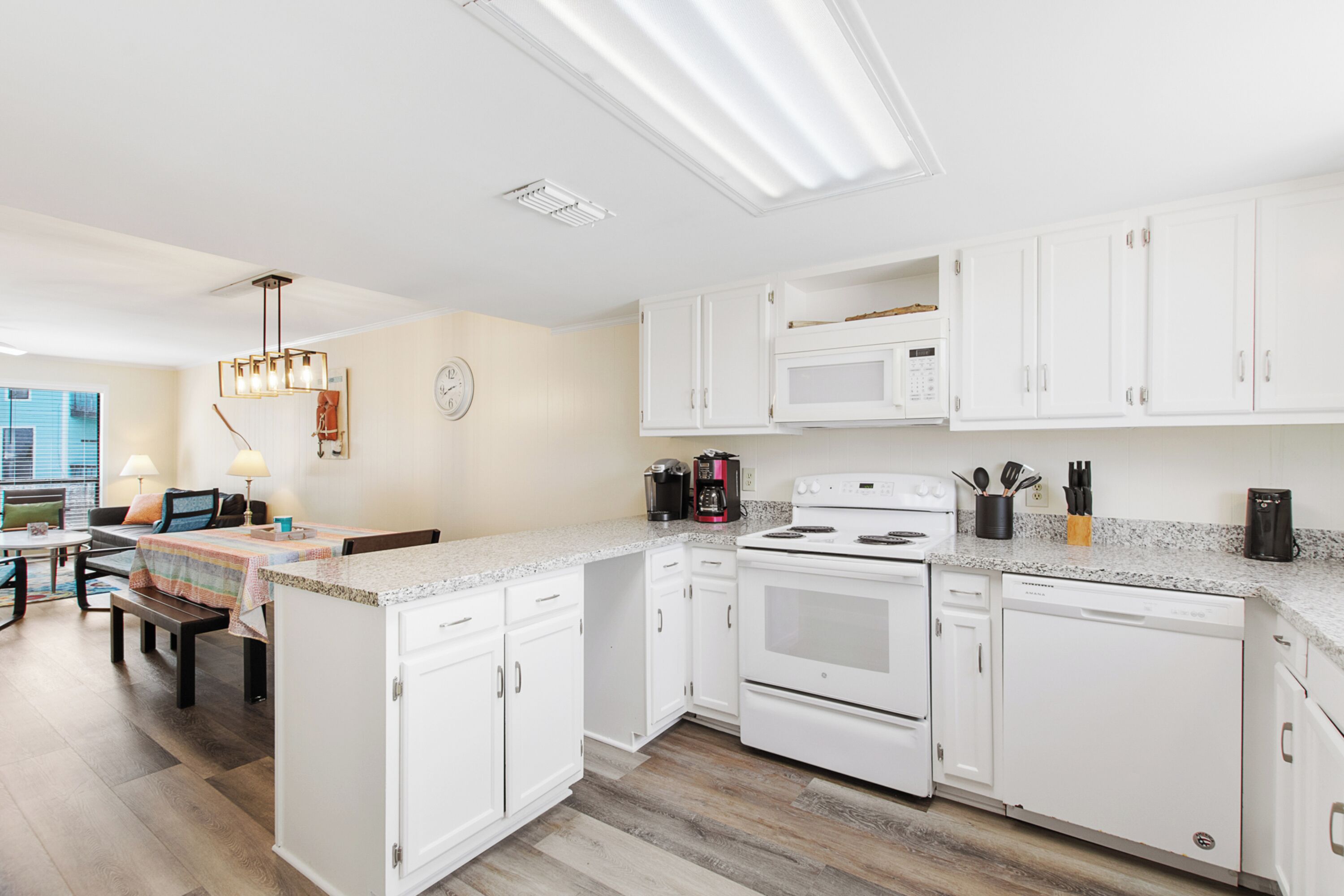 A modern kitchen with white cabinets, a built-in oven, microwave, and dishwasher. There is a dining area with a table and chairs, and a window providing natural light.