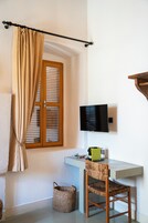 Quarto com porta de madeira, cortinas bege, televisão na parede, escrivaninha com cadeira e cesto no chão.