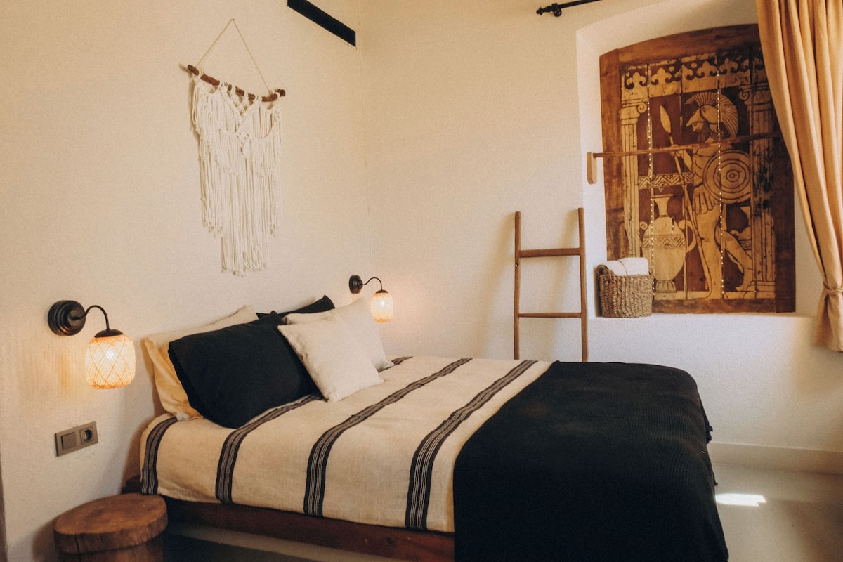 Uma cama com colcha listrada, um pendente de macramé e uma luminária fixada na parede.