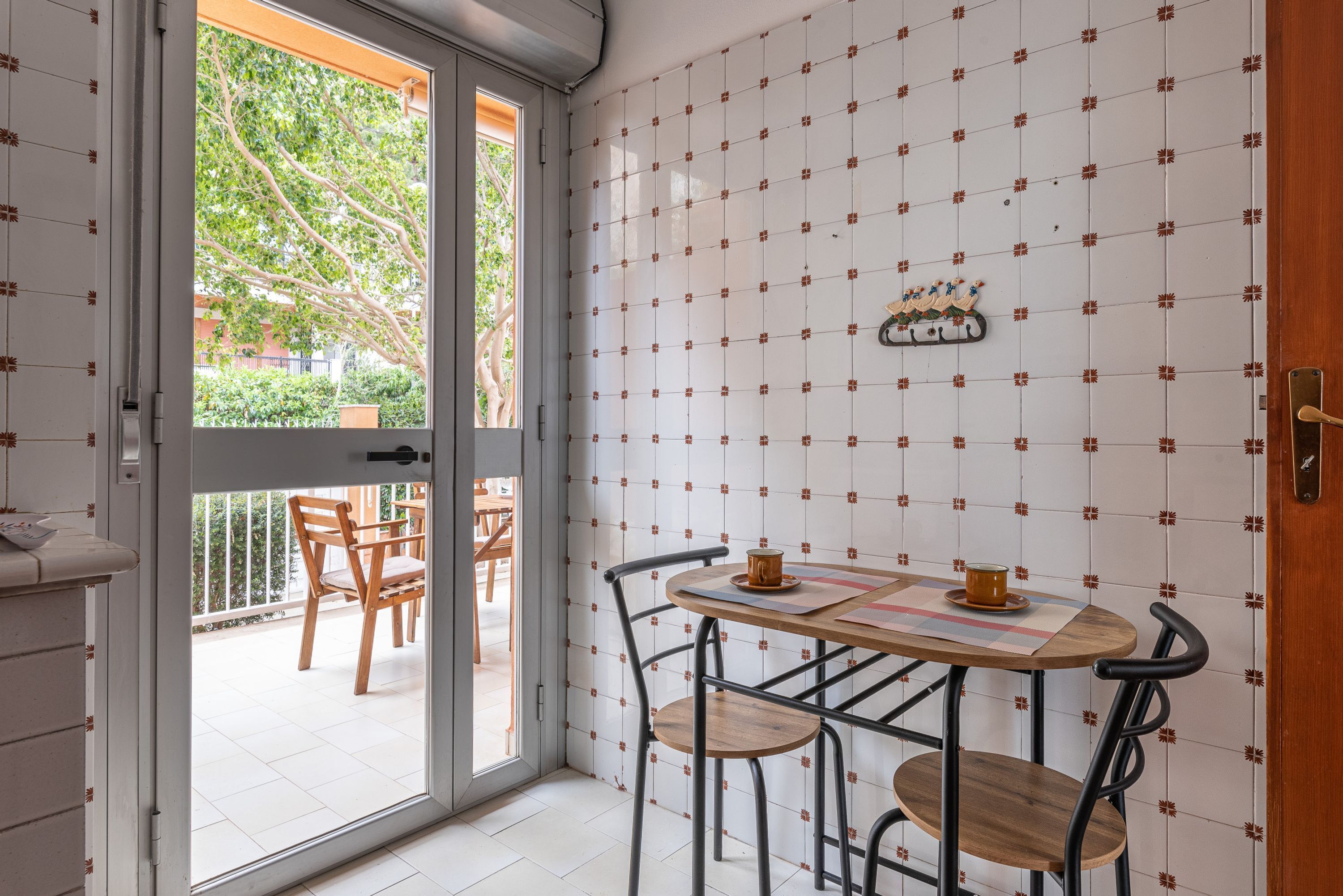A dining area with a table and chairs, a view of the outdoors, and a tiled wall with a decorative pattern.