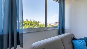 A room with a large window, a sofa, and a view of greenery and buildings. - Addaura Seaview Apartment by Wonderful Italy (Palermo)