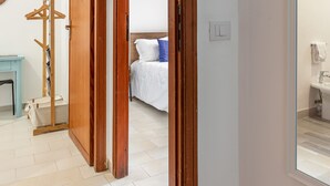 A hotel room with a wooden door, a bed with white bedding, a blue and white striped blanket, a wooden coat rack, and a small table with a chair. - Addaura Seaview Apartment by Wonderful Italy (Palermo)