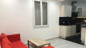 Una sala de estar moderna con un sofá rojo, una mesita de madera y una cocina con armarios de colores claros y electrodomésticos negros.