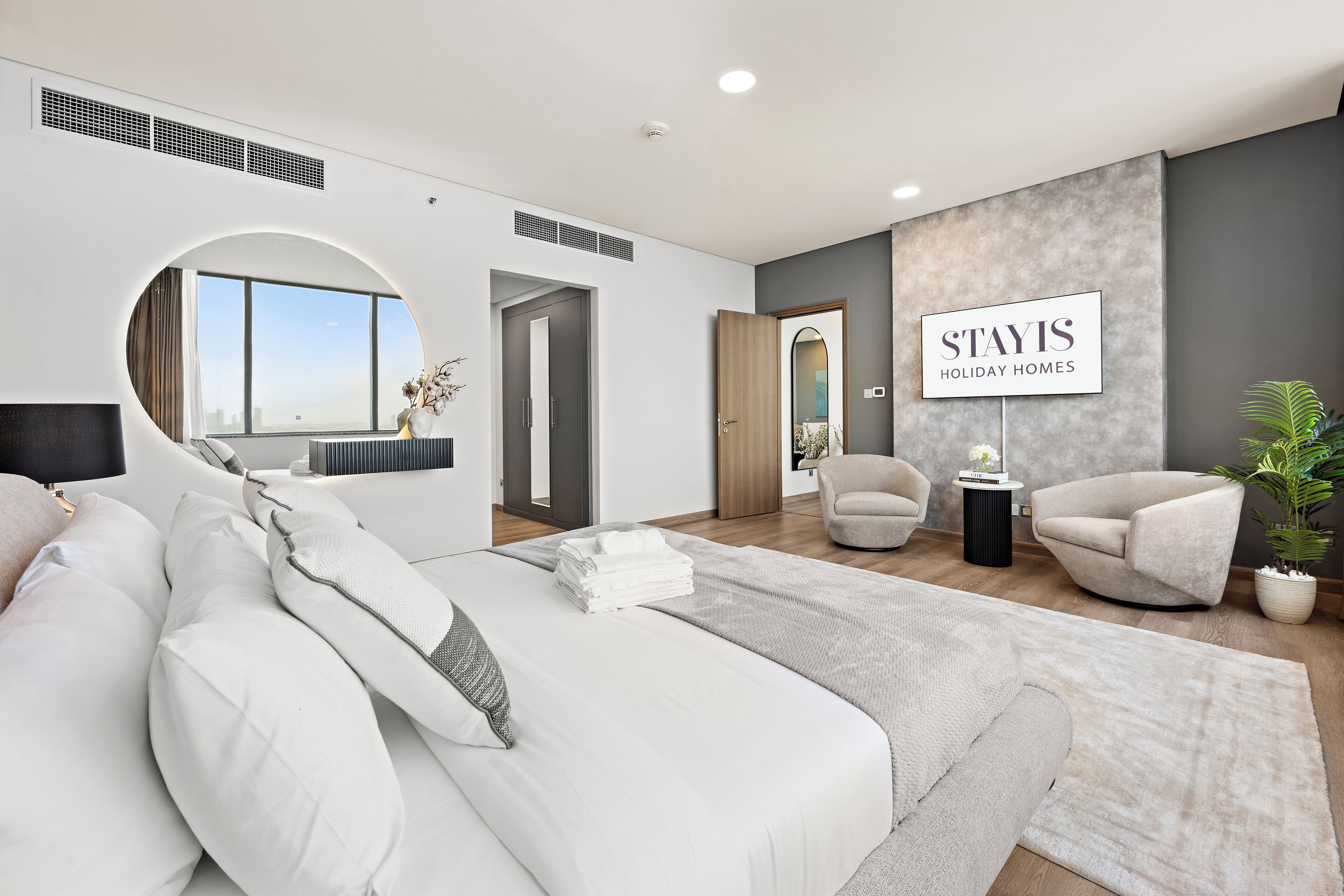 Une chambre d’hôtel moderne avec un grand lit, une fenêtre ronde, une vue sur la ville et une enseigne indiquant « STAYIS HOLIDAY HOMES ».