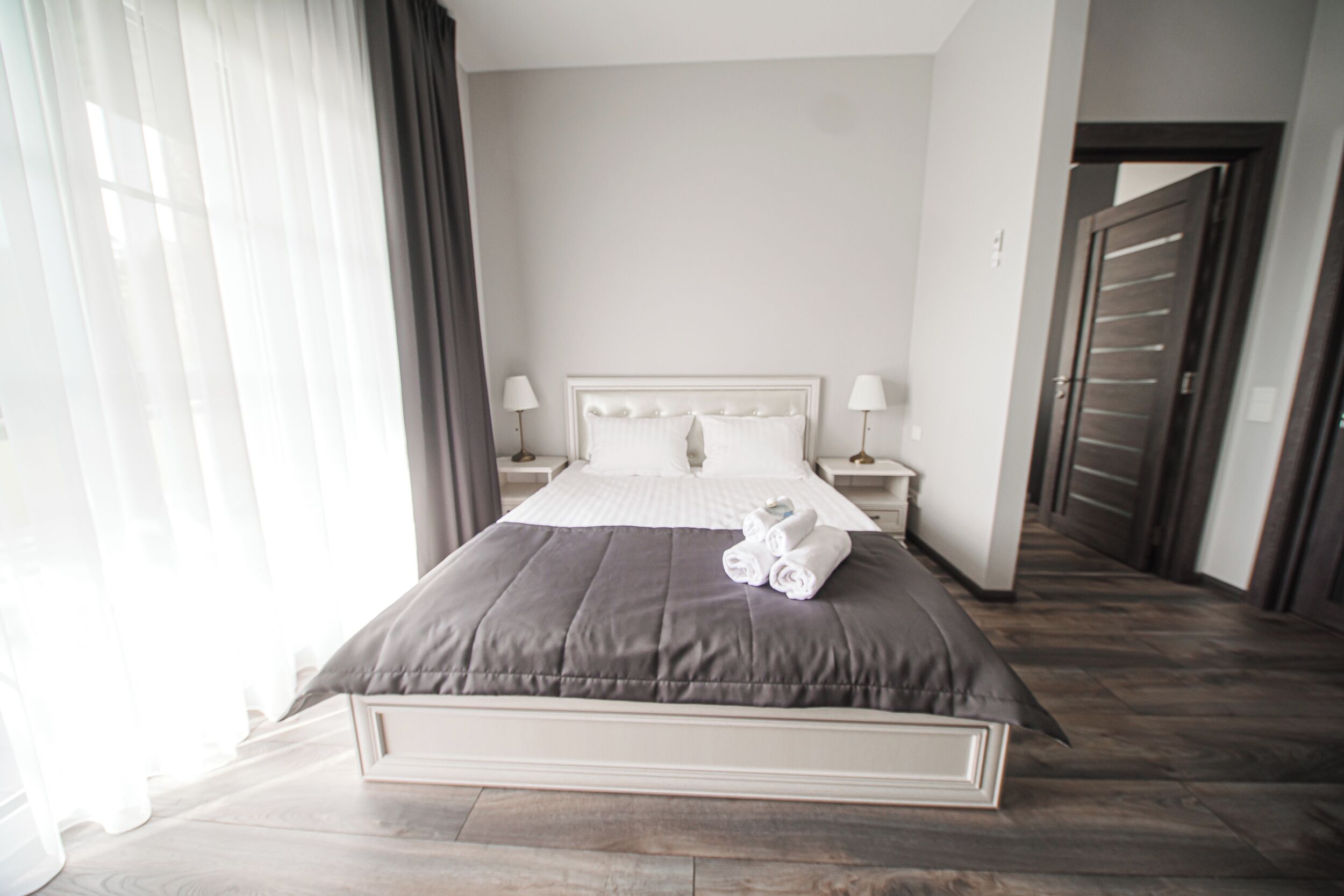A neatly made bed with a grey bedspread and white pillows, two bedside lamps, and a pair of rolled towels on the bed.