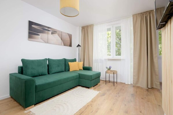 A modern living room with a green sectional sofa, a small side table, and a large window with curtains. - Spacious Apartment for 6 Persons | Balcony | Workspace (Warszawa)