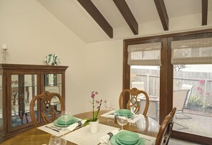 Une salle à manger avec une table en bois dressée pour quatre personnes, un buffet en verre et une vue sur le jardin à travers de grandes fenêtres.
