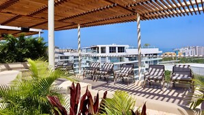 Um lounge no terraço com espreguiçadeiras e uma pérgola de madeira, com vista para a paisagem urbana e o mar.
