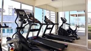 Uma academia com esteiras e bicicletas ergométricas, janelas amplas e um aparelho de ar condicionado.