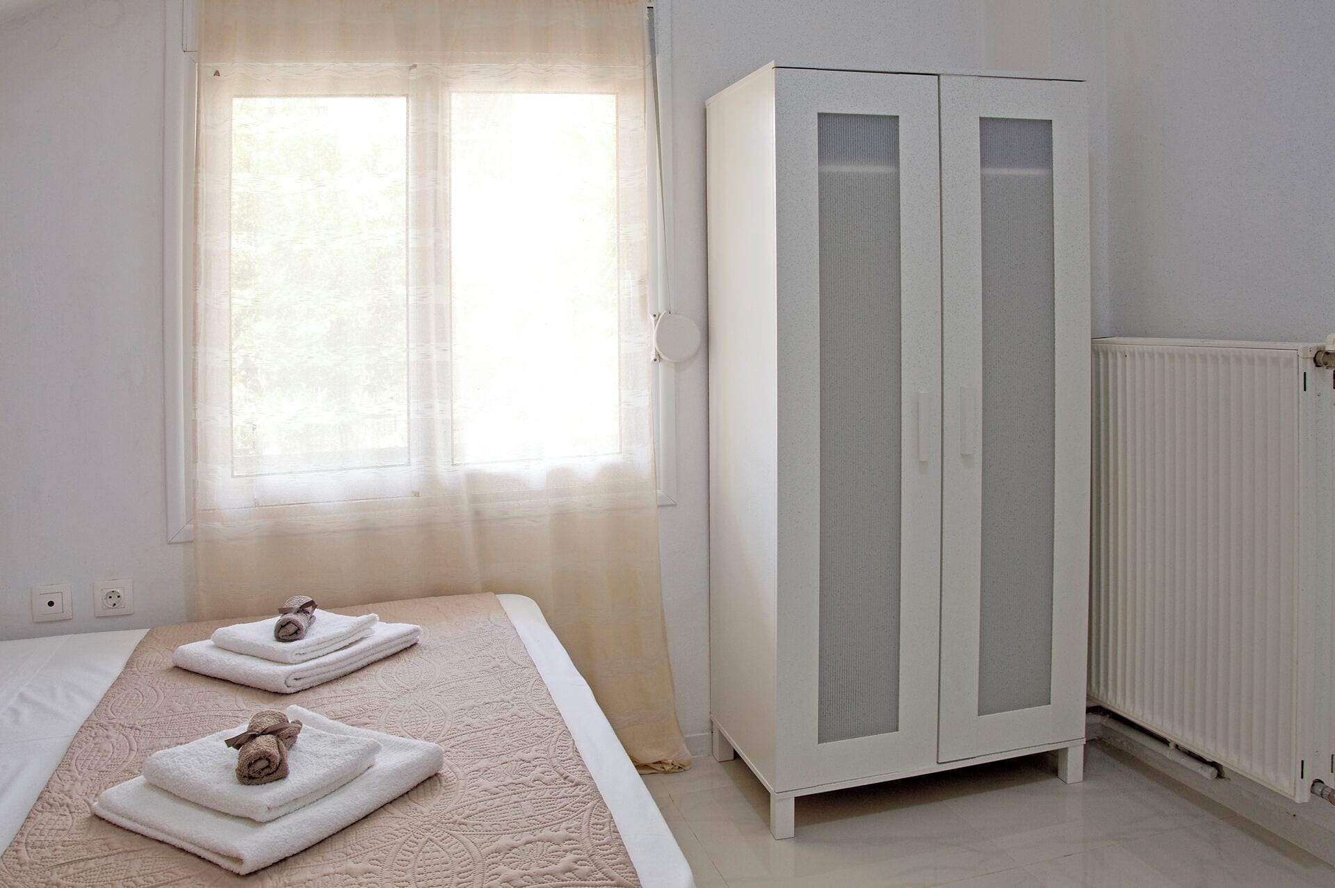 Une chambre à coucher avec un lit, deux serviettes, une armoire, une fenêtre et un radiateur.