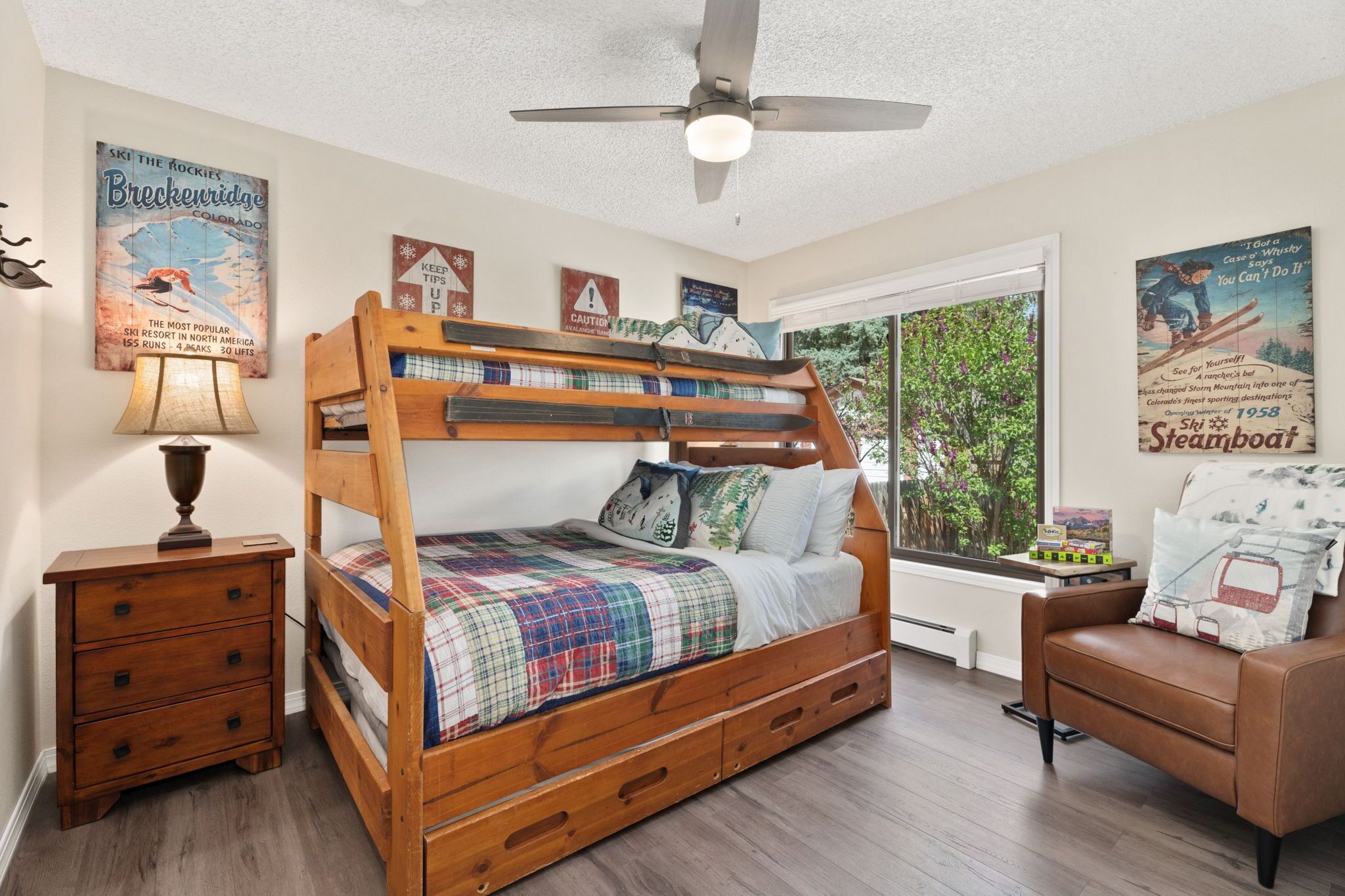 A bedroom with a wooden bunk bed, a nightstand, a lamp, a window with a view of trees, and wall decorations.