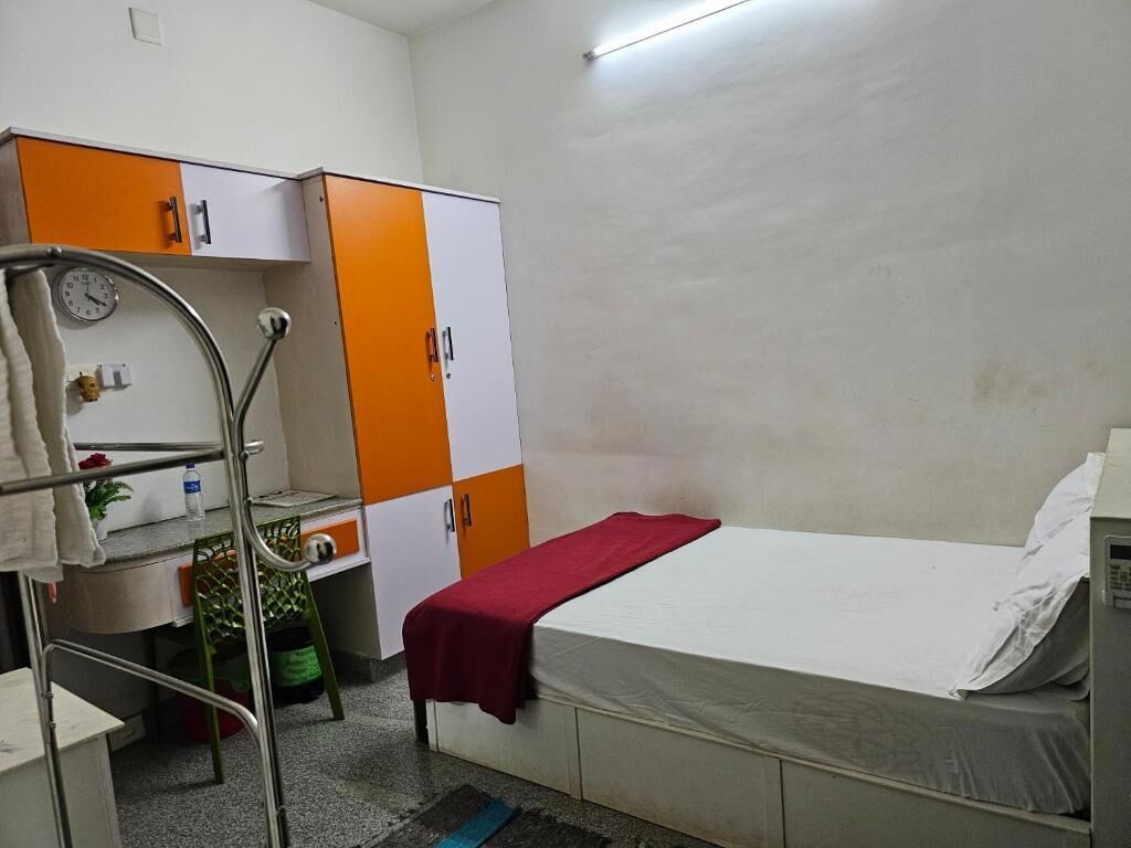 Een kleine eenpersoonskamer met een bed, nachtkastje, stoel, kledingkast en een klok aan de muur.