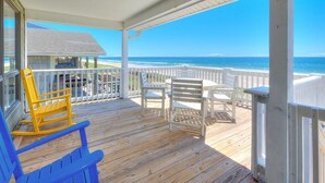 Une terrasse en bois avec des fauteuils Adirondack blancs et jaunes, donnant sur une plage.