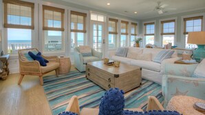 A spacious living room with a large window overlooking the ocean, a ceiling fan, and a variety of seating options. - Discover beachfront bliss at Starboard Side, Oak Island's spacious 4-bedroom retreat. (Oak Island)