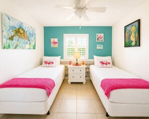 A bedroom with two beds, a window with blinds, a ceiling fan, and artwork on the walls. - Flamingo Flats Ocean Pointe 5306 2BR/2BA (Tavernier)