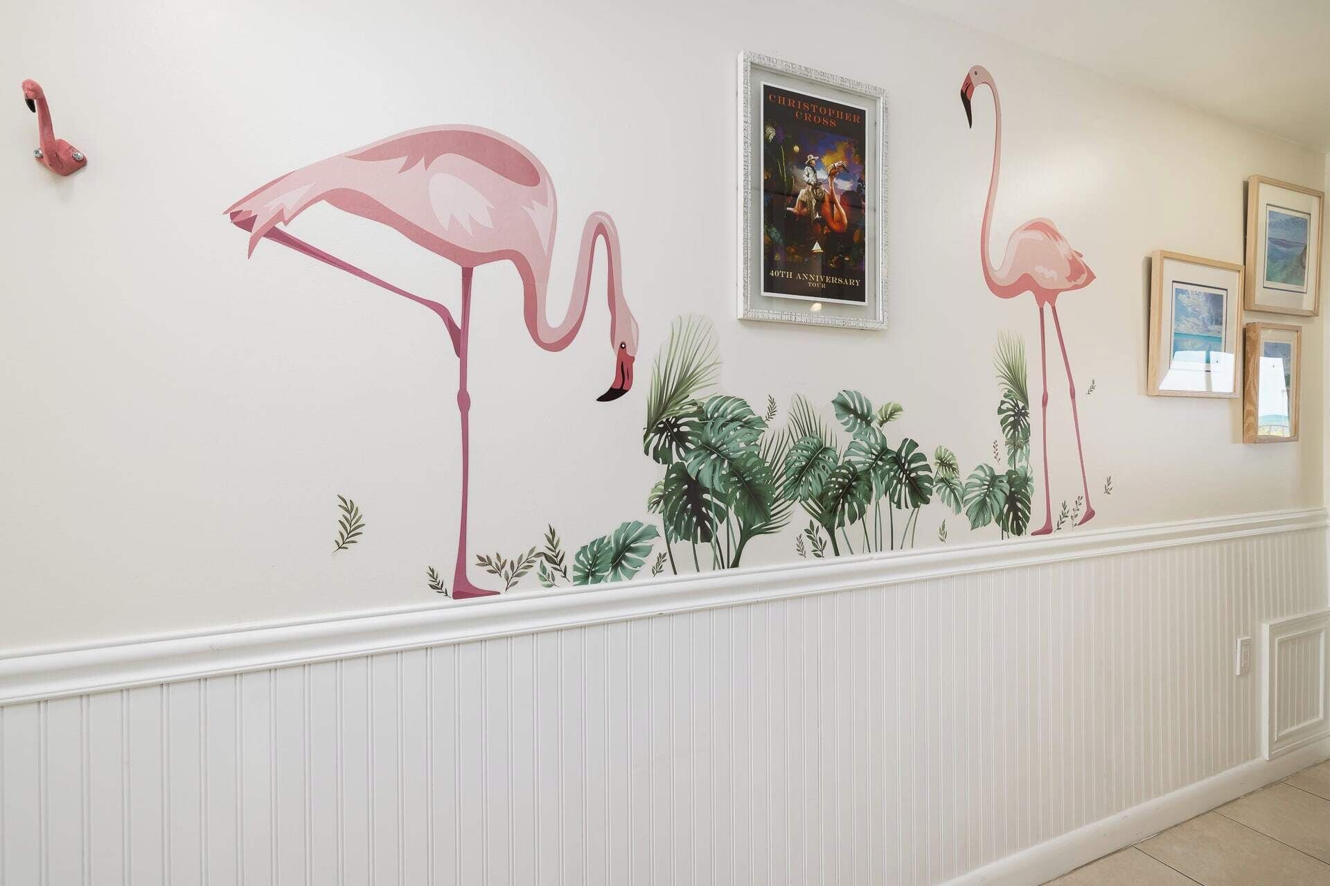 Un pasillo con un mural de flamencos y follaje tropical, cuadros enmarcados y paneles de madera blanca.