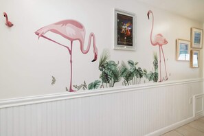 A hallway with a wall mural of flamingos and tropical foliage, framed artwork, and a white wainscoting. - Flamingo Flats | Marina View | Ocean Pointe 5306 (Tavernier)