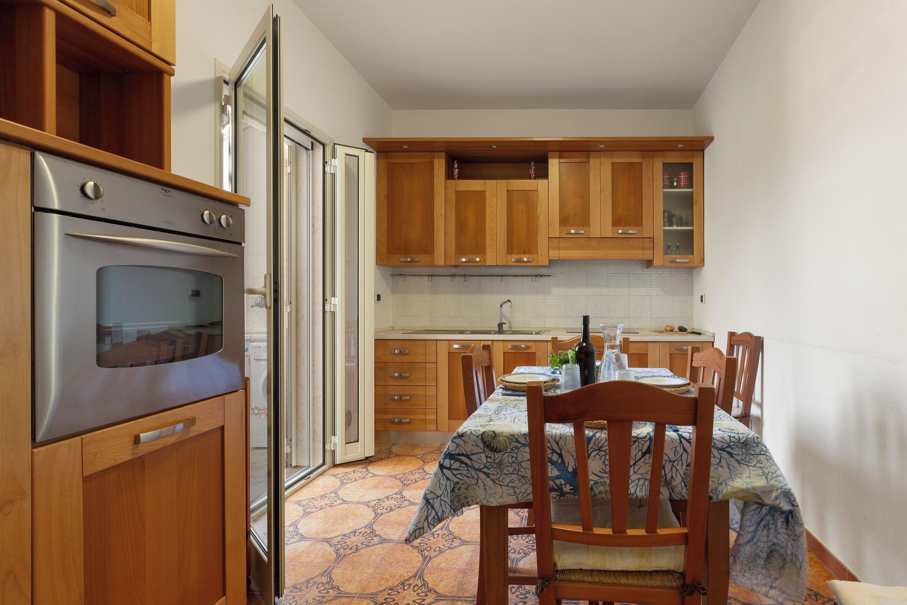 A compact kitchen with wooden cabinets, a built-in oven, a dining table with chairs, and a tiled backsplash.