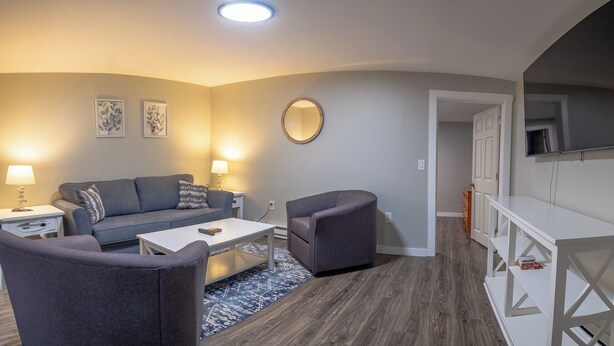 A modern living room with a grey sofa, two armchairs, a coffee table, and a TV mounted on the wall. - Beautiful Apartments in Thorsen Road Maine (Hancock)