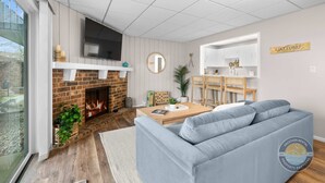 Ein modernes Wohnzimmer mit Kamin, einem grauen Sofa, einem Holztisch und einer Küchenzeile mit Barbereich.