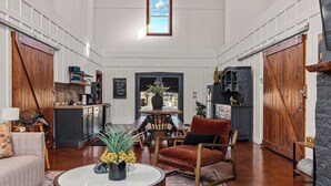 Amplia sala de estar con piso de madera, una mesa de centro redonda con un florero, un sillón rojo y un sofá. Cuenta con una zona de cocina con ventana sobre el fregadero y un comedor con mesa y sillas.
