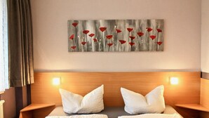 Ett hotellrum med en sänggavel i trä, två vita kuddar, en tavla med röda blommor på väggen och två sänglampor.