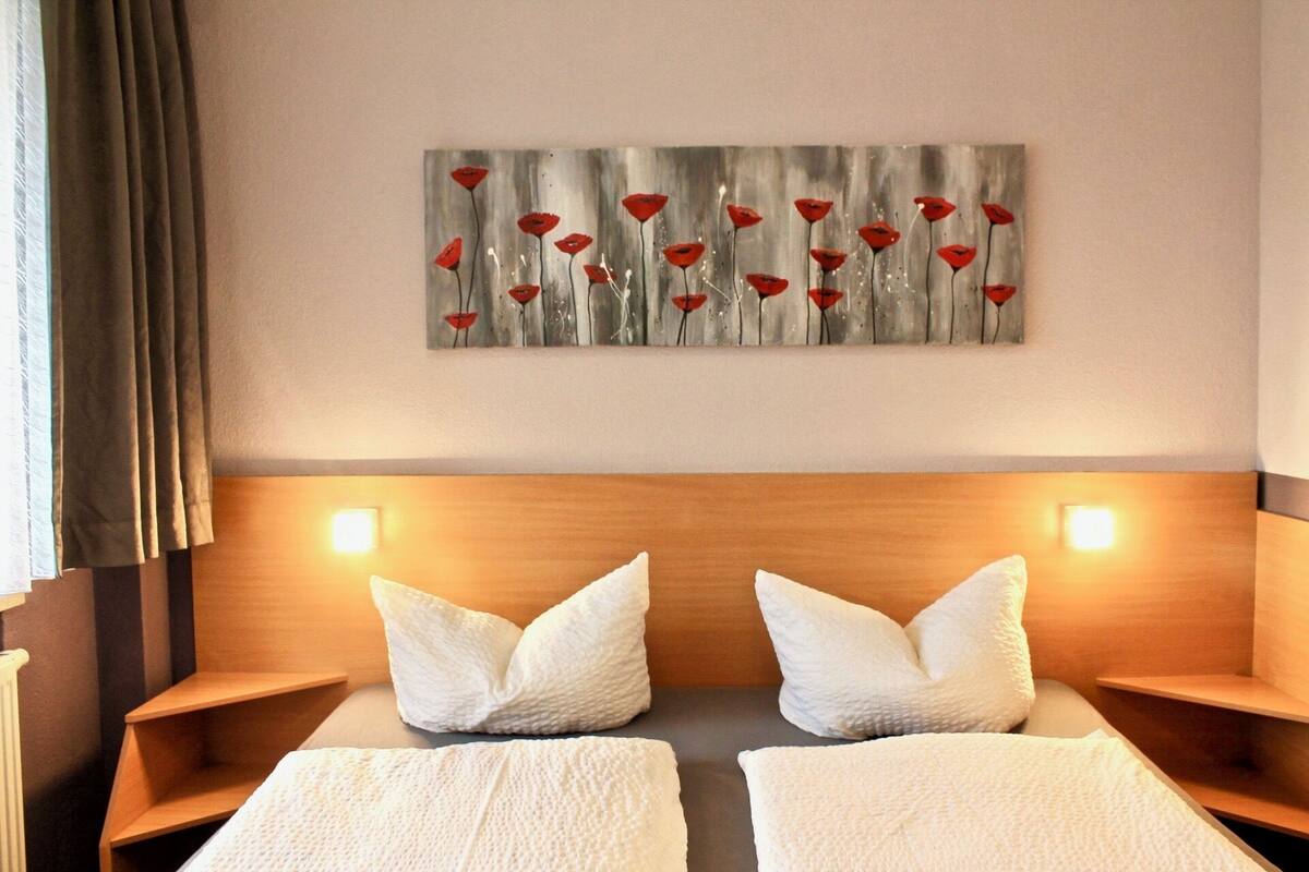 A hotel room with a wooden headboard, two white pillows, a painting of red flowers on the wall, and two bedside lamps.
