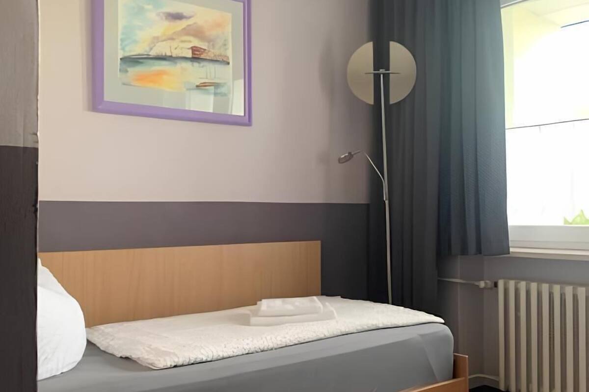 A neatly arranged bedroom with a bed, a bedside table, a floor lamp, a window with curtains, and a painting on the wall.