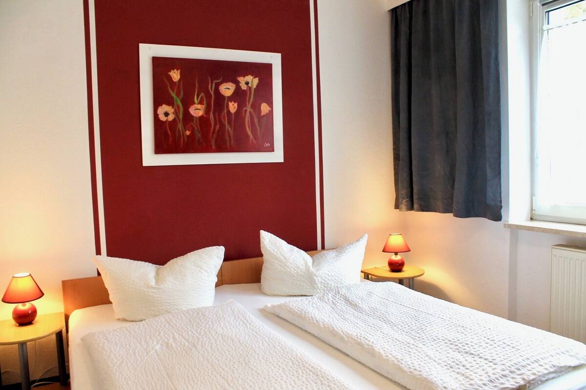 A hotel room with a bed, white pillows, a red accent wall, a framed floral painting, two bedside lamps, and a window with curtains.