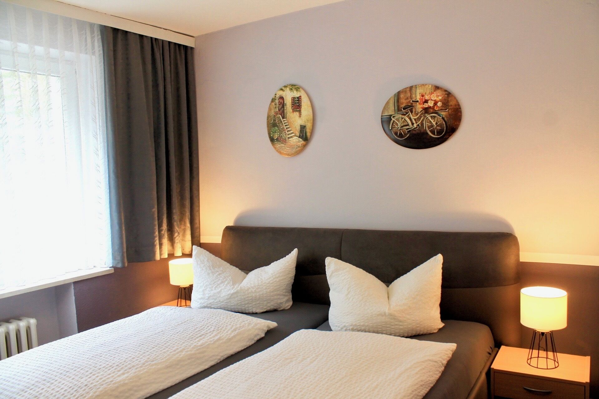 A hotel room with a bed, two wall art pieces, a window with curtains, and a bedside lamp.