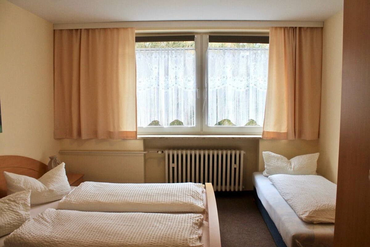 A room with two beds, beige curtains, a window with a view, and a radiator.