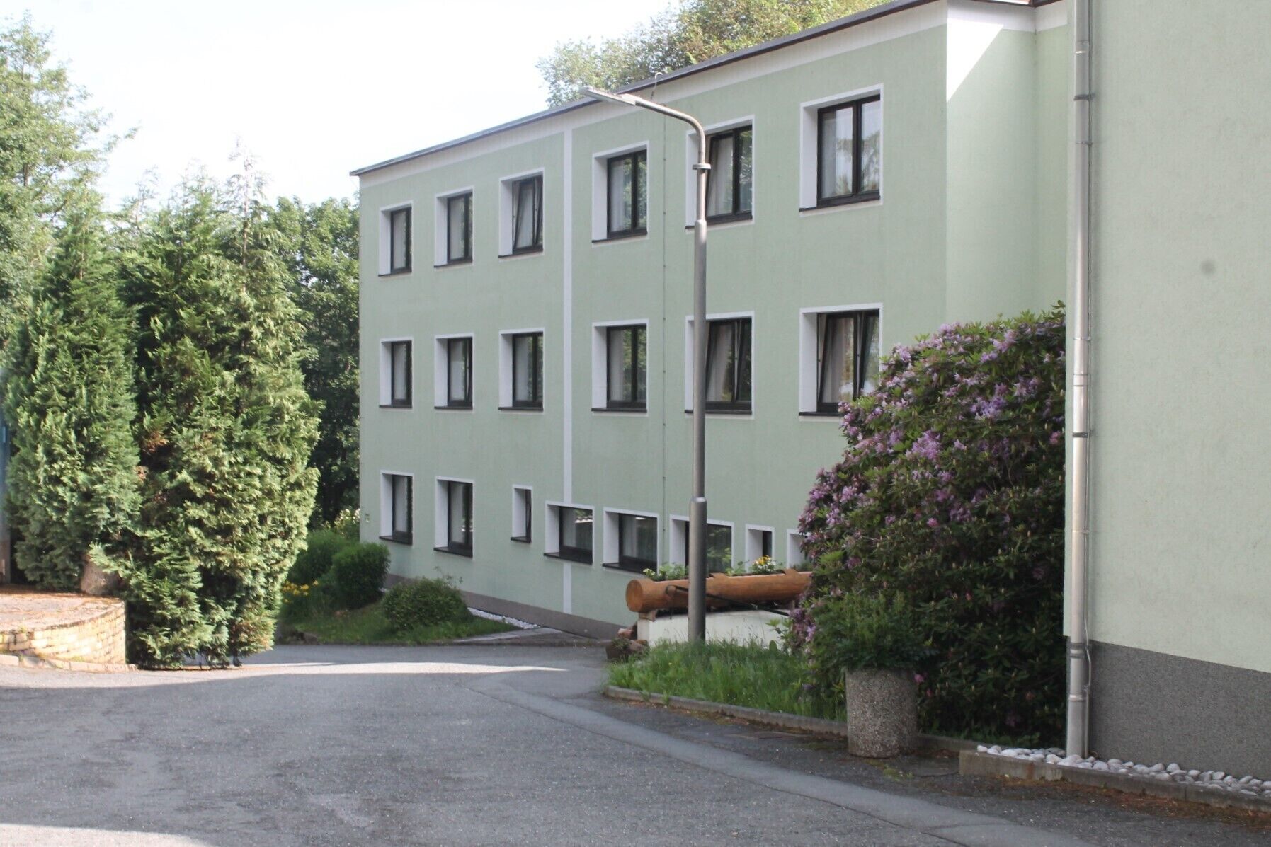 Un edificio de varios pisos con fachada verde claro, múltiples ventanas y un pequeño jardín con arbustos y flores.