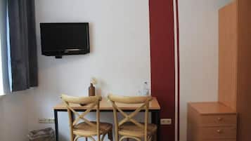A room with a desk, two chairs, a television mounted on the wall, a small cabinet, and a trash bin.
