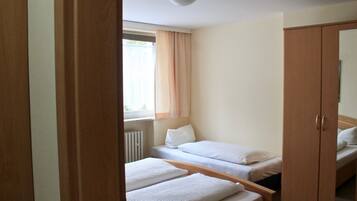 A small, compact room with two single beds, a wooden wardrobe, and a window with curtains.
