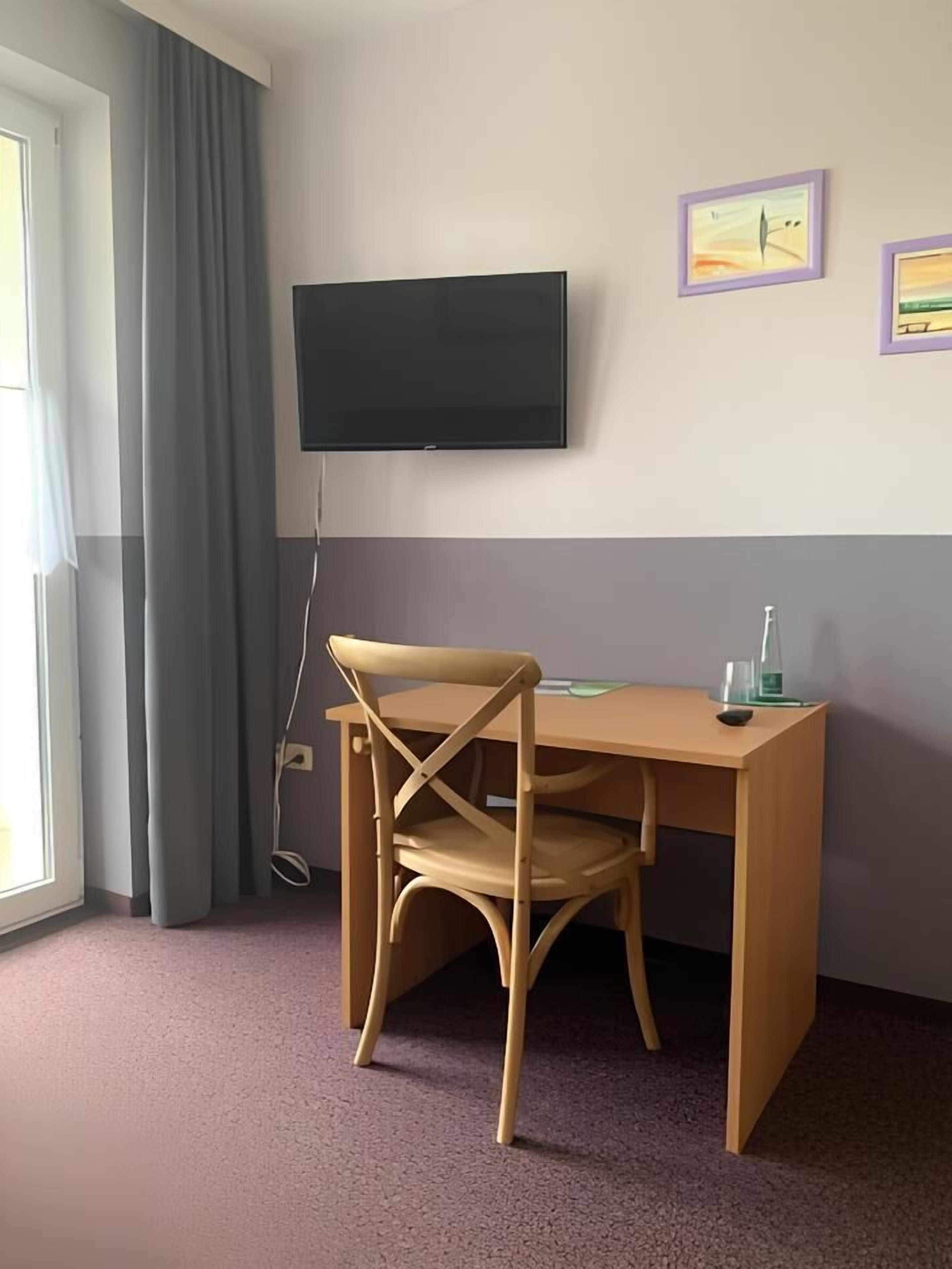A room with a wooden desk, a chair, a flat-screen TV mounted on the wall, and a window with curtains.