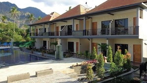 Een hotel met twee verdiepingen, een zwembad, terras buiten en een bergachtig landschap op de achtergrond.