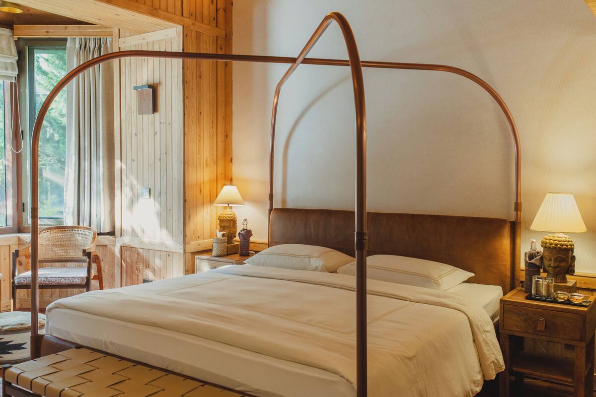 Un lit à baldaquin avec une tête de lit en bois, deux tables de chevet avec des lampes, un fauteuil en osier et une vue sur l’extérieur par une fenêtre.
