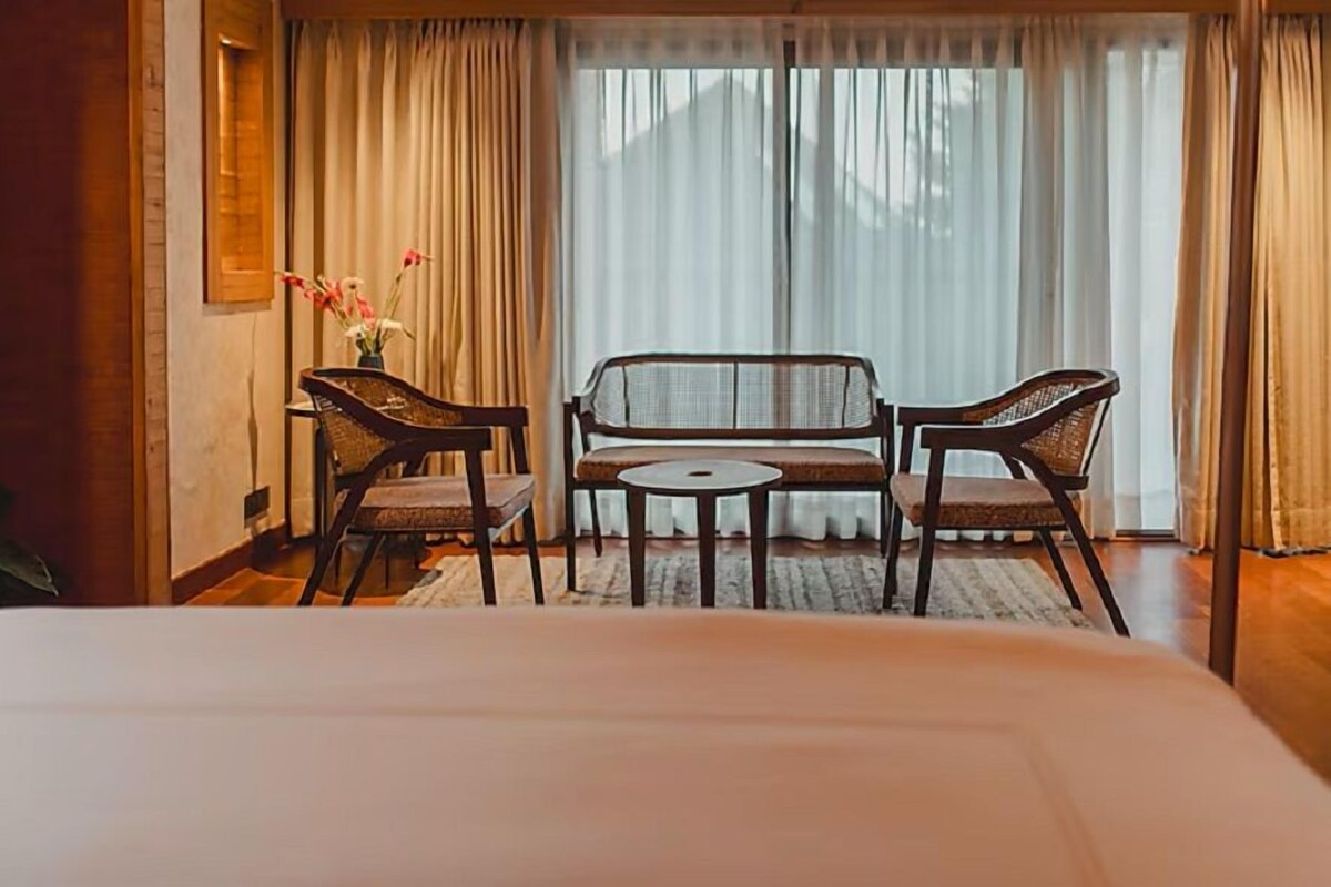 A hotel room with a bed, a small table, two wicker chairs, a vase with flowers, and a view of the outdoors through sheer curtains.