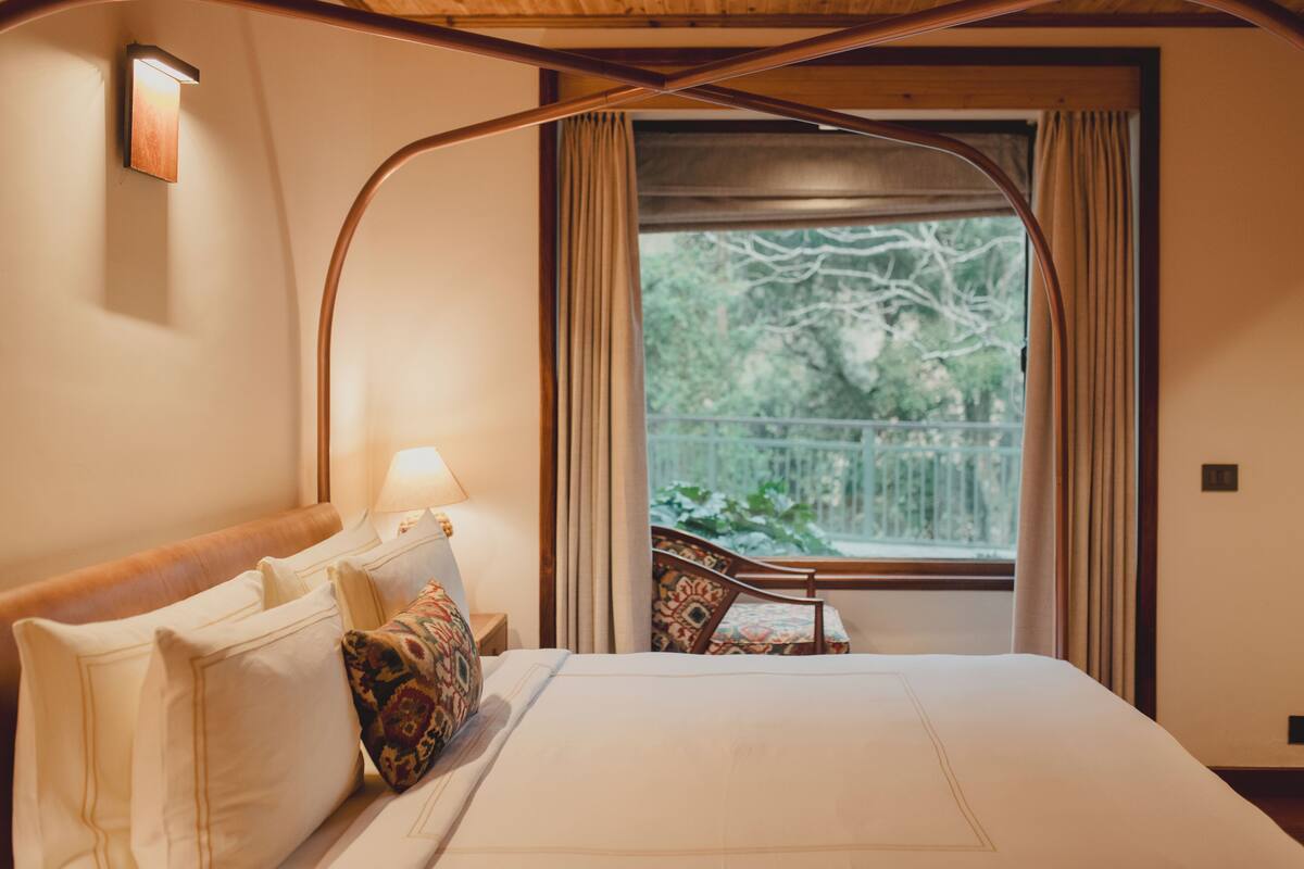 Une chambre d’hôtel avec un grand lit, une table de chevet avec une lampe, une fenêtre avec des rideaux et une vue sur la verdure.
