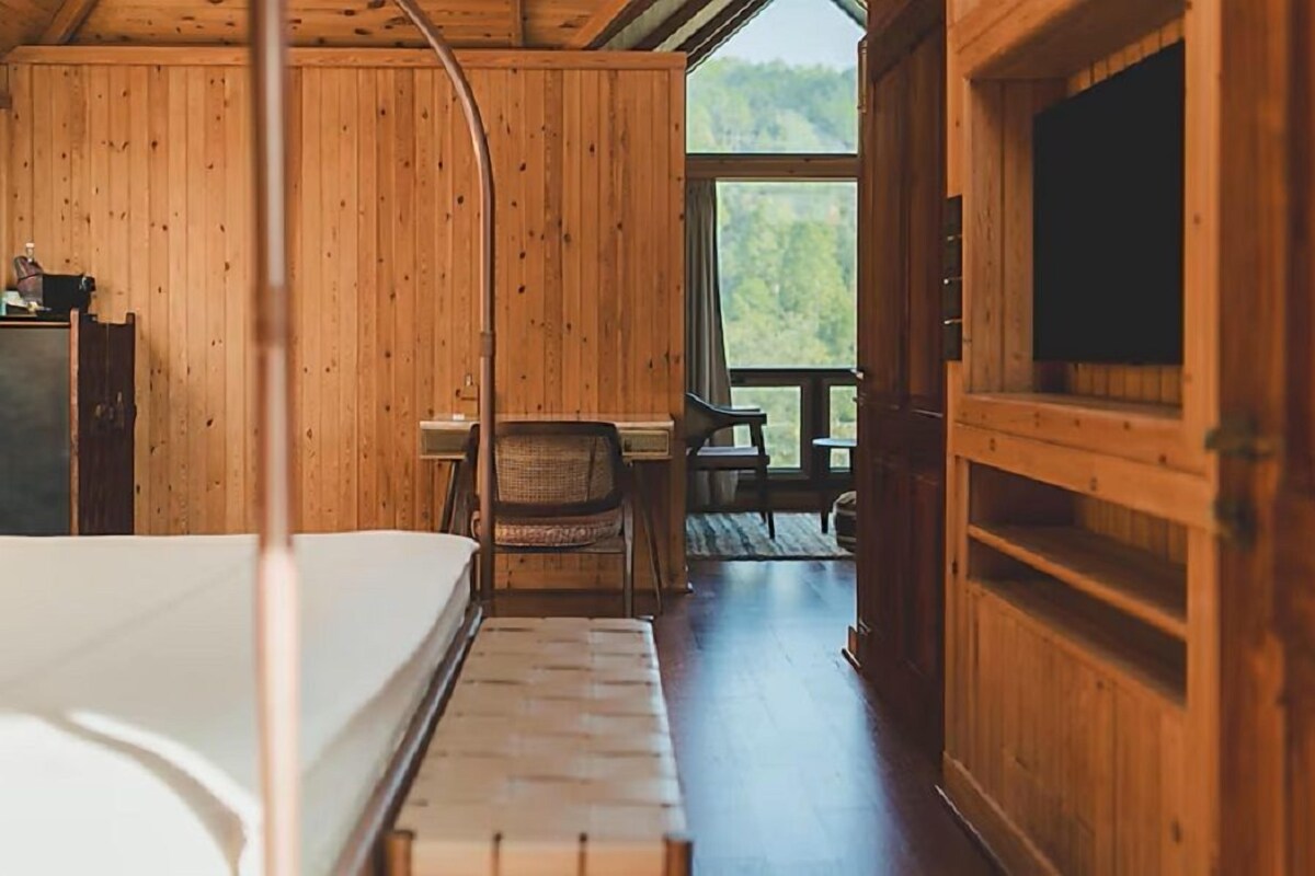 A wooden cabin interior with a bed, a chair, a desk, and a large window offering a view of the outdoors.