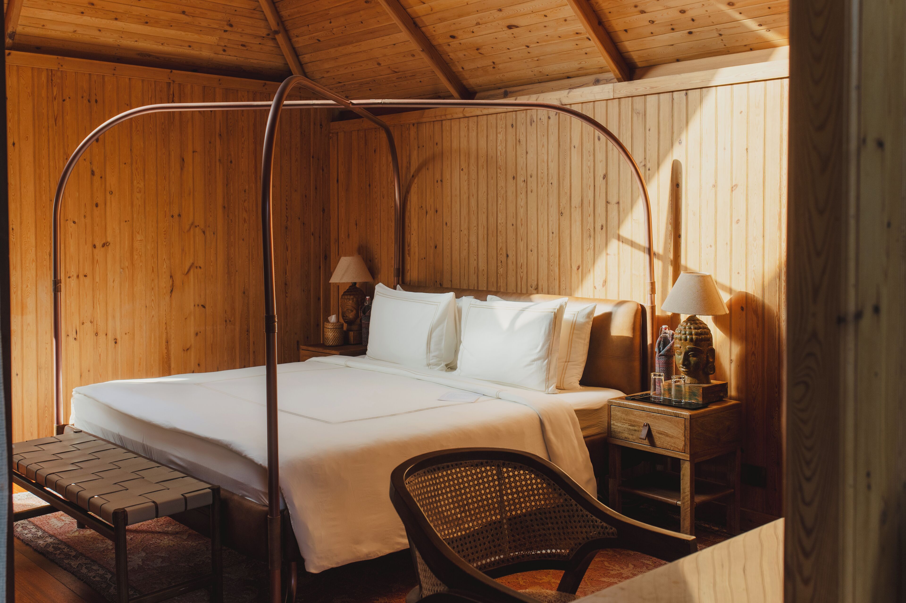 A wooden cabin bedroom with a four-poster bed, a nightstand with a lamp, and a wicker chair.