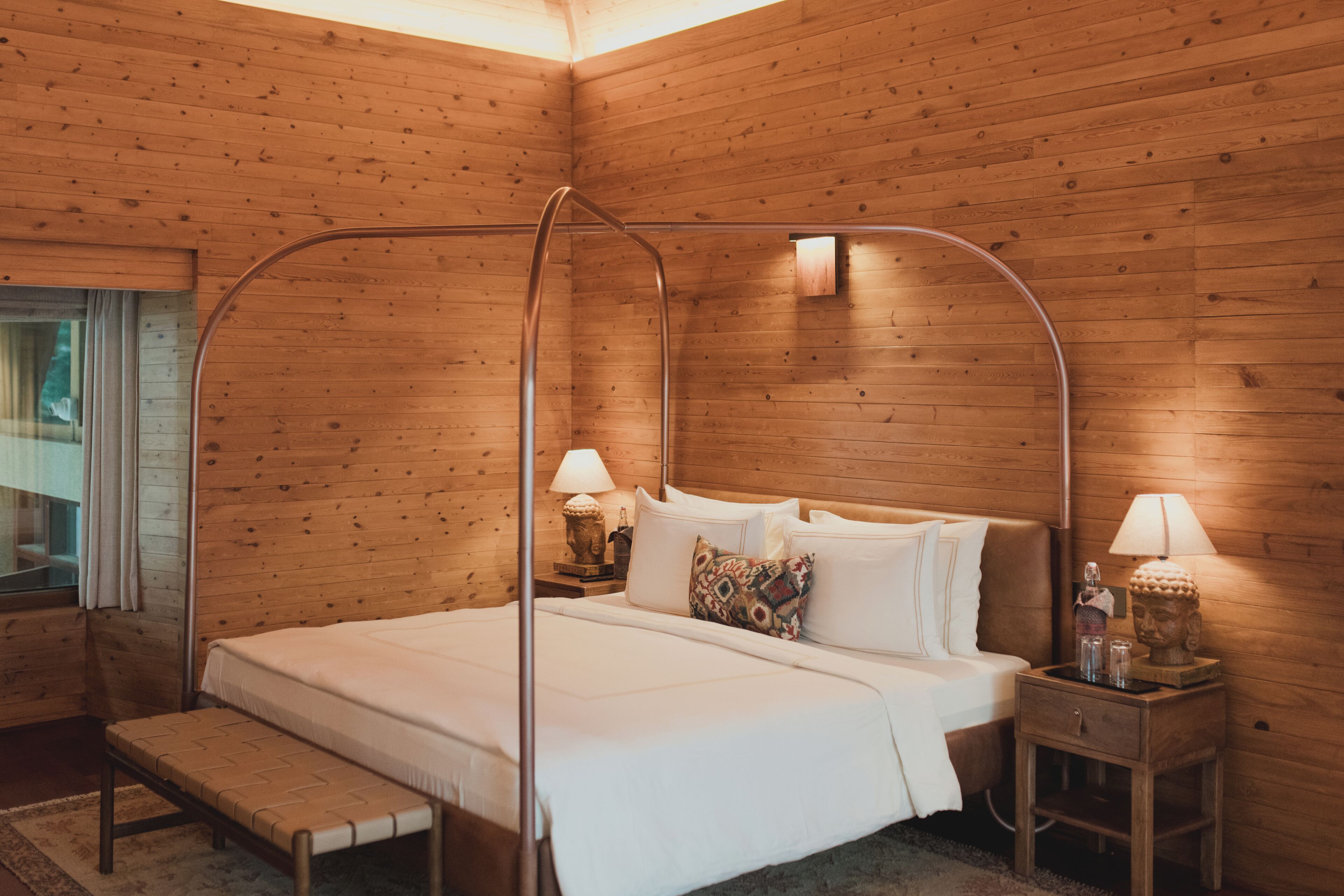 Una habitación con paneles de madera, una cama, dos mesitas de noche con lámparas, un banco y una ventana con cortinas.